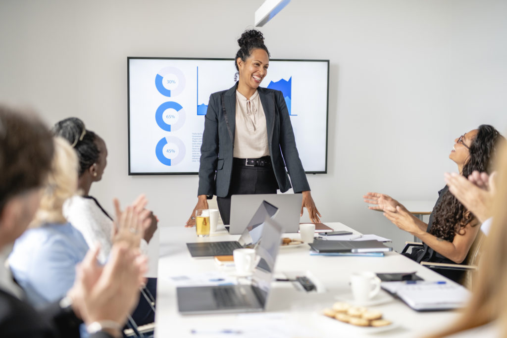 Personal perspective of Hispanic business professional in early 40s smiling as colleagues applaud successful presentation in office board room.