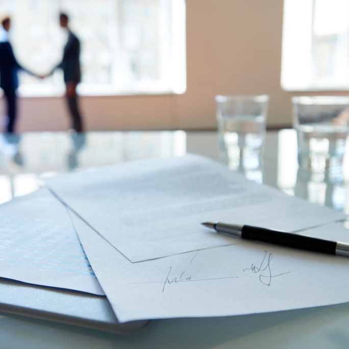 Signed business contract lying on table, business partners shaking hands in the background